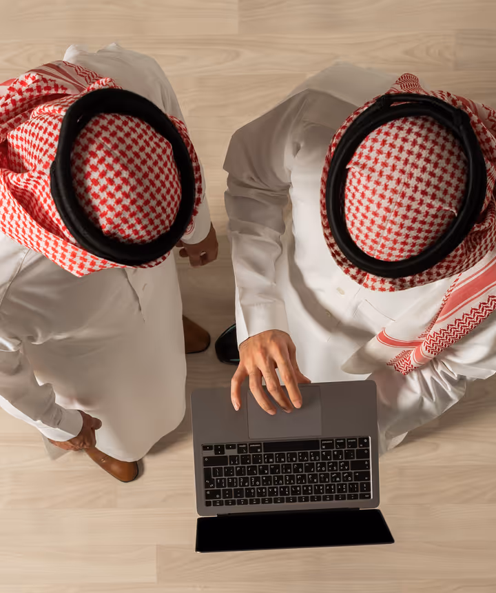Two men in traditional Saudi dress working together at a laptop, photographed from above