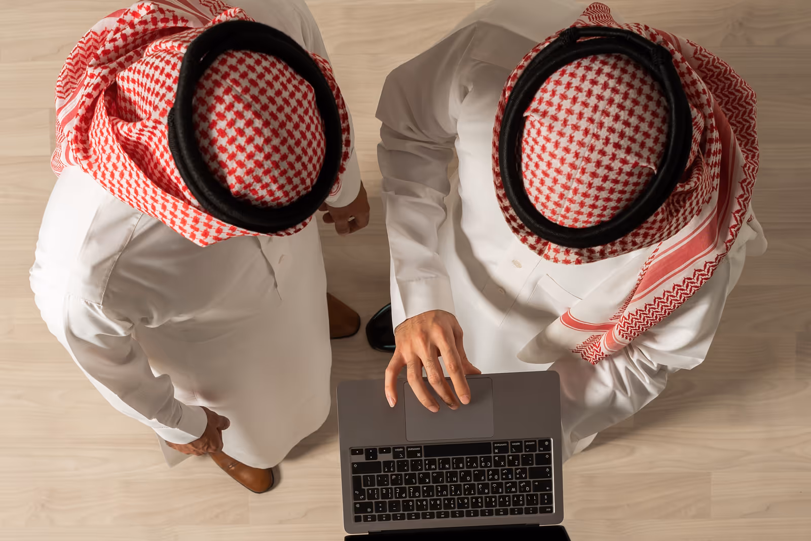 رجلان بالزي السعودي التقليدي يعملان معًا على حاسب محمول، بلقطة من الأعلى